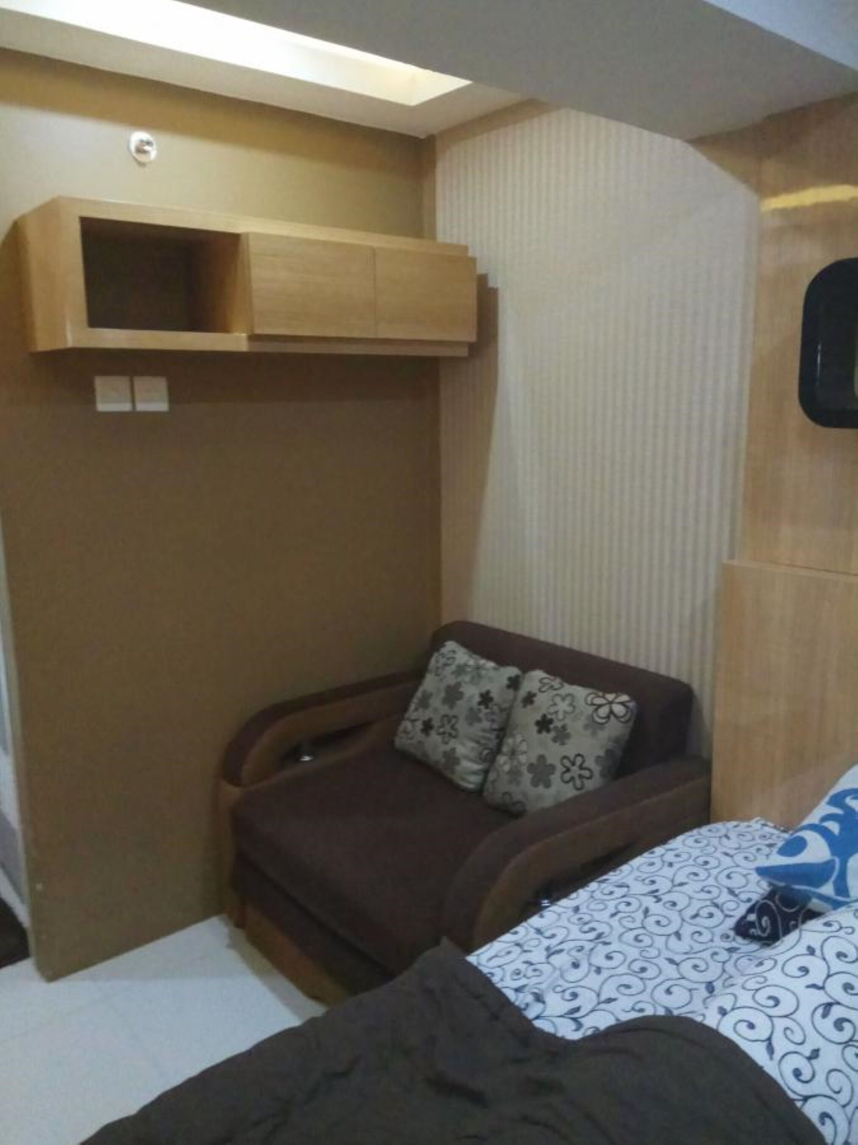 A compact room with a bed, a small seating area with a chair and cushions, and a wooden shelf unit.