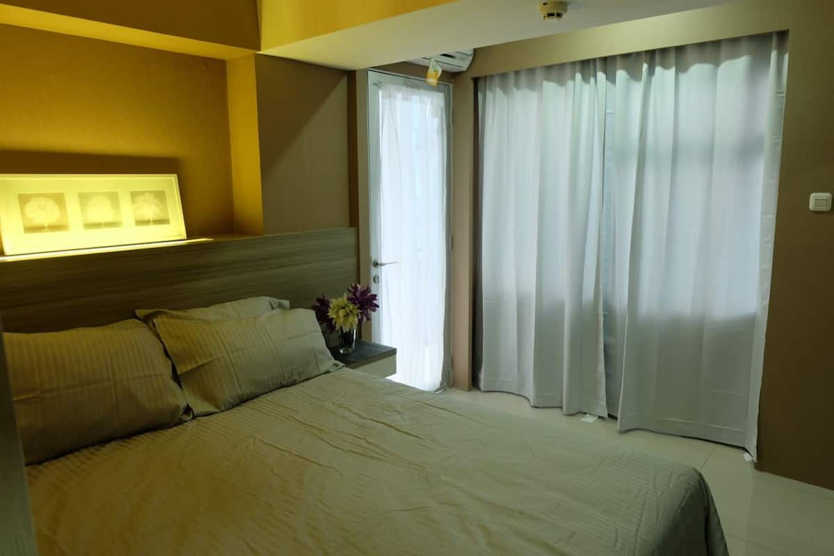 A hotel room with a bed, a nightstand with a vase of flowers, a window with white curtains, and a wall-mounted light fixture.