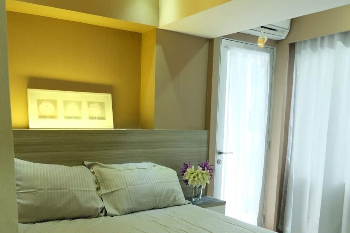 A hotel room with a bed, a bedside table with a vase of flowers, a window with curtains, and a wall-mounted air conditioning unit.