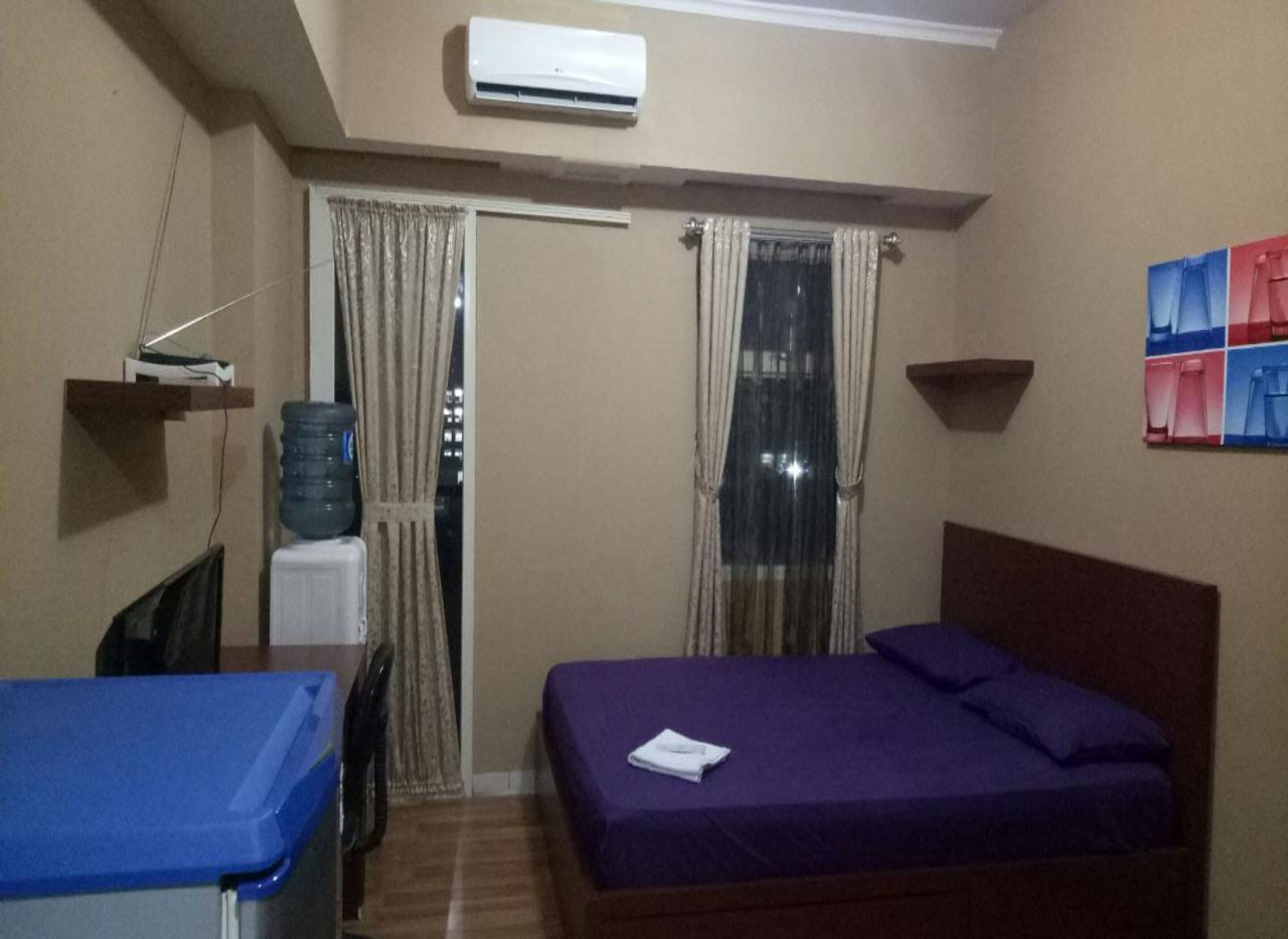 A small hotel room with a bed, a desk, a chair, a water dispenser, and a window with curtains.