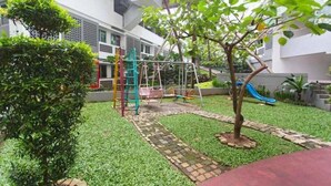 Un patio con un parque infantil, que incluye un tobogán y estructuras para escalar, rodeado de follaje y un camino pavimentado.