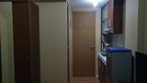 Un pasillo estrecho con un armario de madera, una puerta cerrada, una pequeña zona de cocina con refrigerador y una alfombra roja en el suelo.