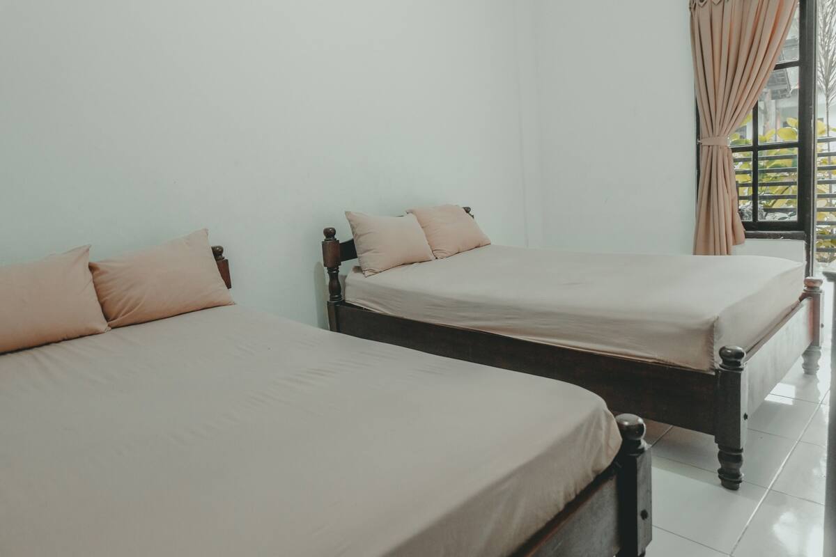 Two single beds with wooden headboards and beige bedding in a room with white walls and a large window with curtains.