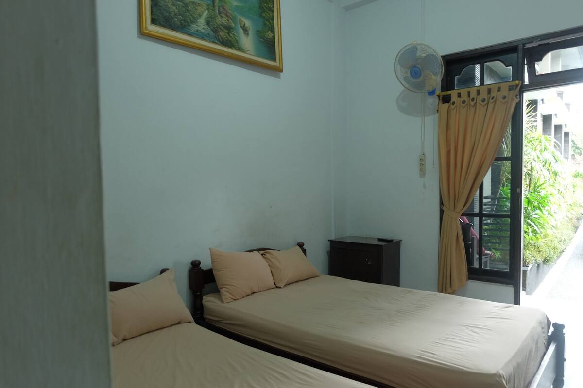 A room with two beds, a painting on the wall, a window with curtains, and a fan.