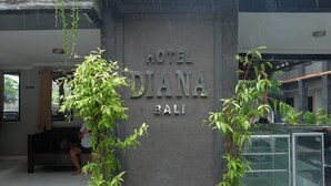 Señal de hotel con el nombre "HOTEL DIANA BALI" y una zona de recepción con recepcionista, plantas en macetas y una vitrina de vidrio.