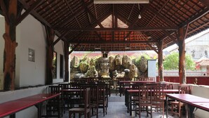 Un espacio al aire libre cubierto con mesas y sillas de madera, suelo de baldosas y vista a un edificio tradicional de techo rojo al fondo.