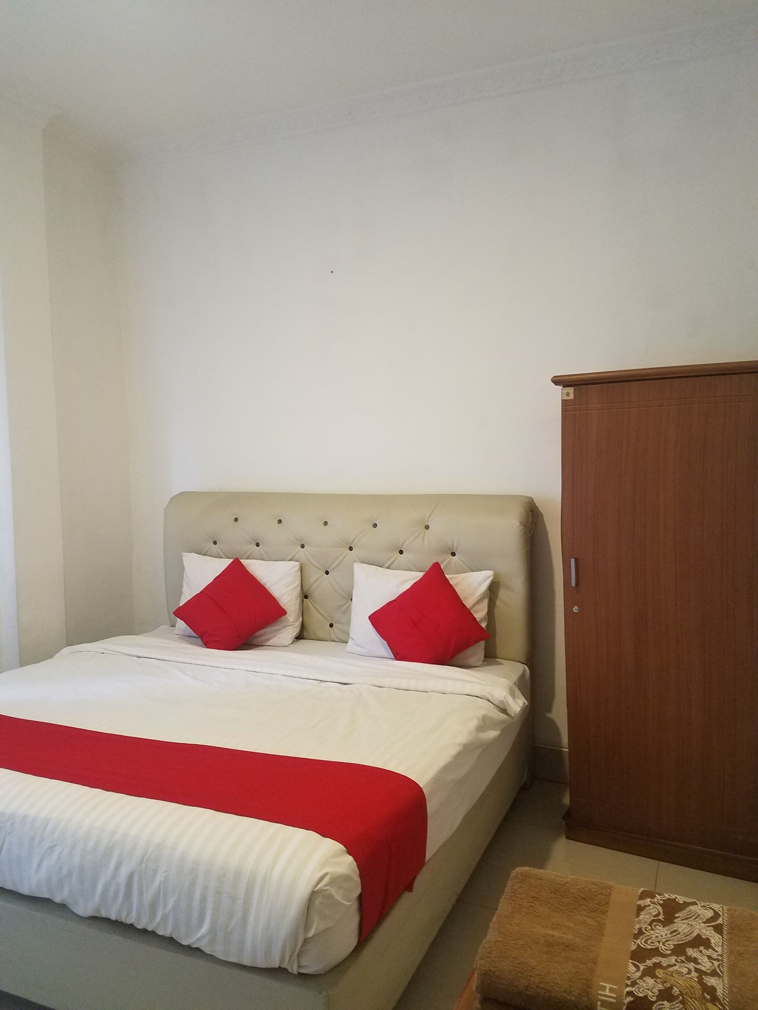 Un lit bien fait, recouvert d’une couvre-lit rouge et blanche, une armoire en bois et un petit pouf marron.