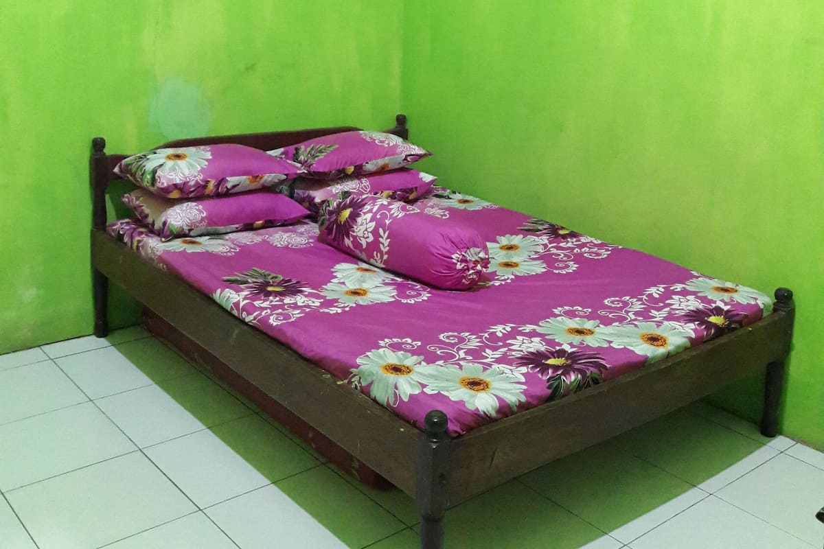 A single bed with a floral bedspread and multiple pillows against a green wall, in a room with white tiled flooring.