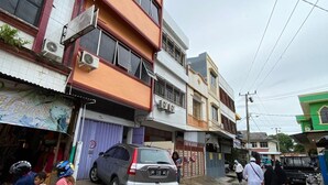 A street scene with buildings, a parked car, and pedestrians. - Kost Aka (Makassar)
