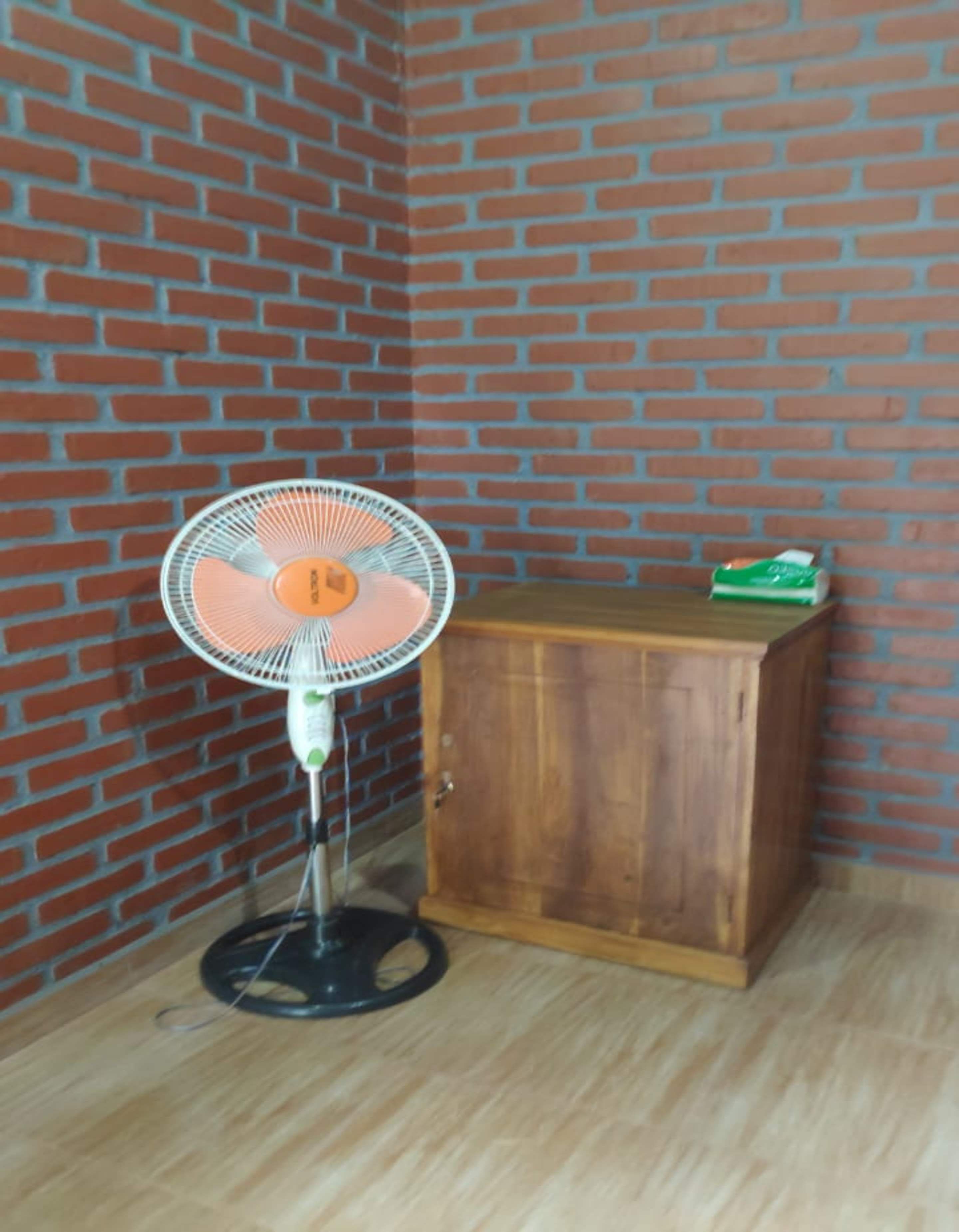 A corner room with a brick wall, a wooden cabinet, and a standing fan.