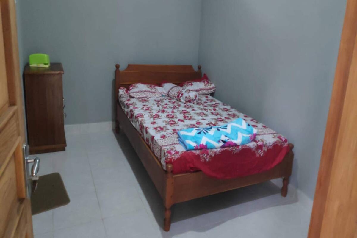 A small, single-bed room with a wooden bed frame, a green cabinet, and a tiled floor.