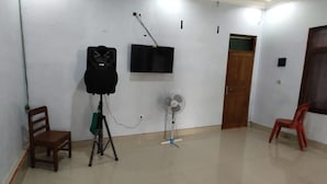 A room with a flat-screen TV mounted on the wall, a tripod with a black object on it, a standing fan, and two wooden chairs. - Wahyu Utomo Syariah (Tawangmangu)