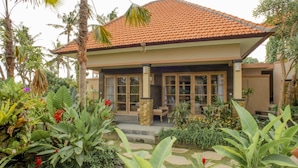 Une maison à deux étages, avec un toit en tuiles, de grandes fenêtres et un jardin planté de plantes tropicales.