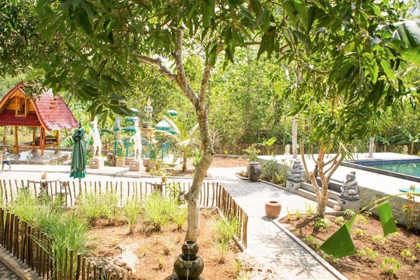 Un jardín con una estructura de juego de madera, un pequeño edificio de techo rojo y una zona de piscina.