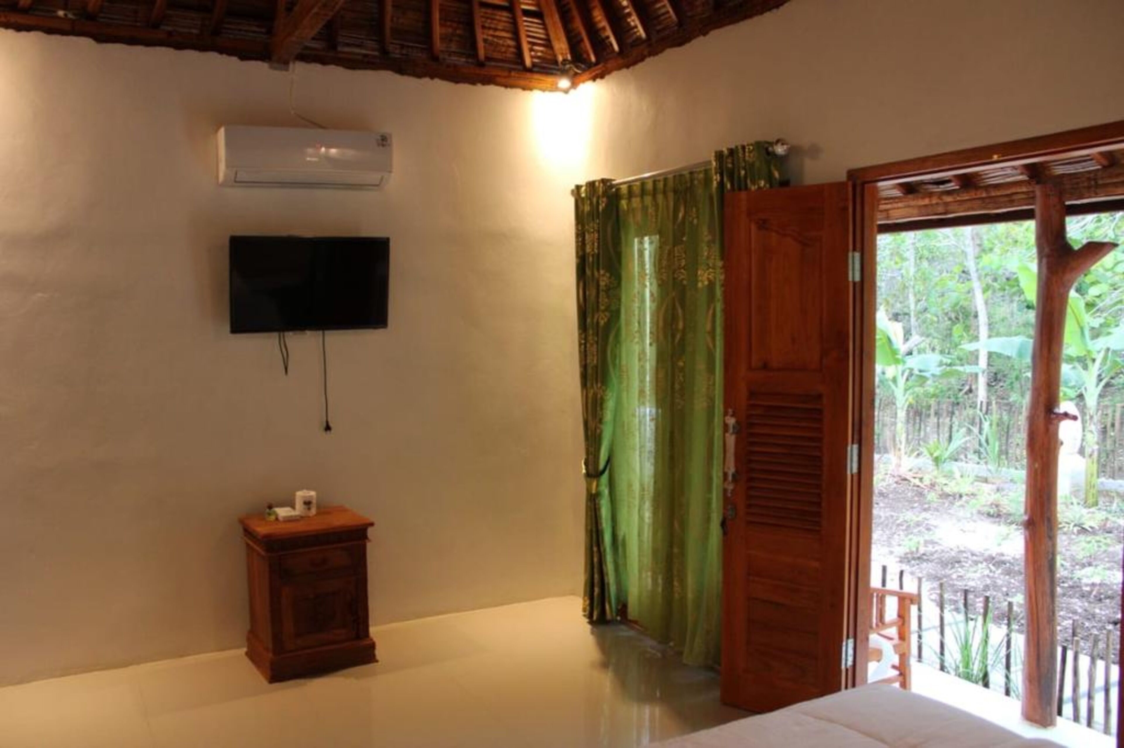 Una habitación con una cama, televisión montada en la pared, una mesita de noche de madera y una ventana con cortinas verdes.