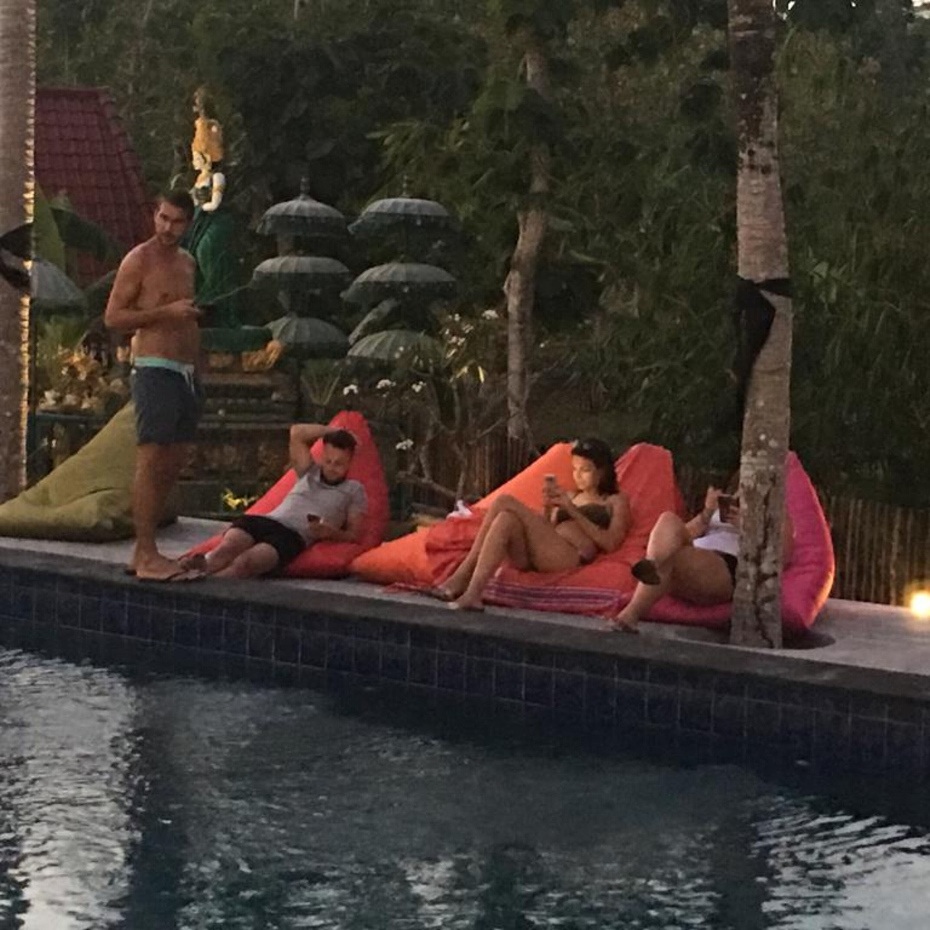 Ein Poolbereich mit gefliestem Rand, eine Person steht daneben, mehrere Personen entspannen auf bunten Kissen.