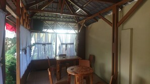 Un espace extérieur abrité, doté d’une table et de chaises, offrant une vue sur la verdure environnante et surmonté d’une structure de toit en métal ondulé.