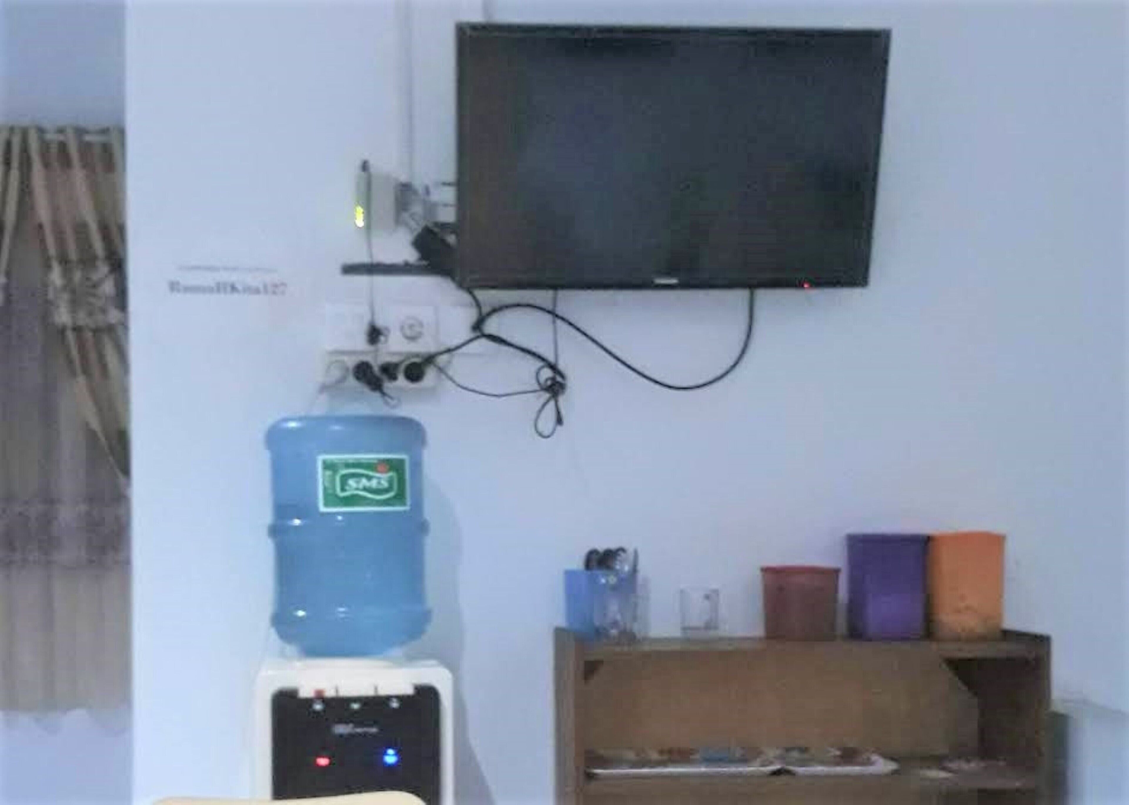 A room with a mounted television, a water dispenser, and a shelf with various items.