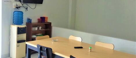 Un salón de conferencias con una larga mesa, cuatro sillas, un televisor en la pared, un dispensador de agua y un estante con diversos objetos.