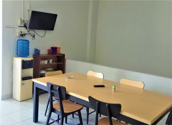 Un salón de conferencias con una larga mesa, cuatro sillas, un televisor en la pared, un dispensador de agua y un estante con diversos objetos.