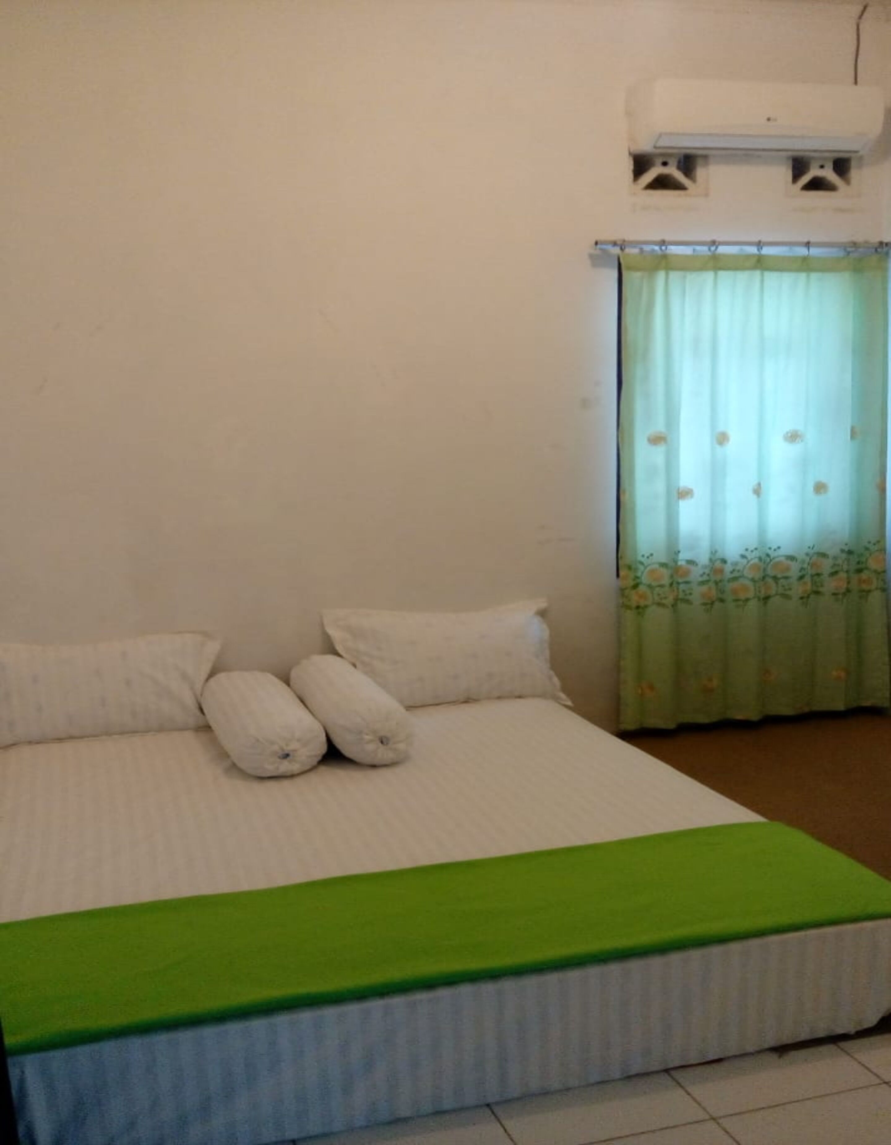 A single bed with a green bedspread, white pillows, and a floral curtain.