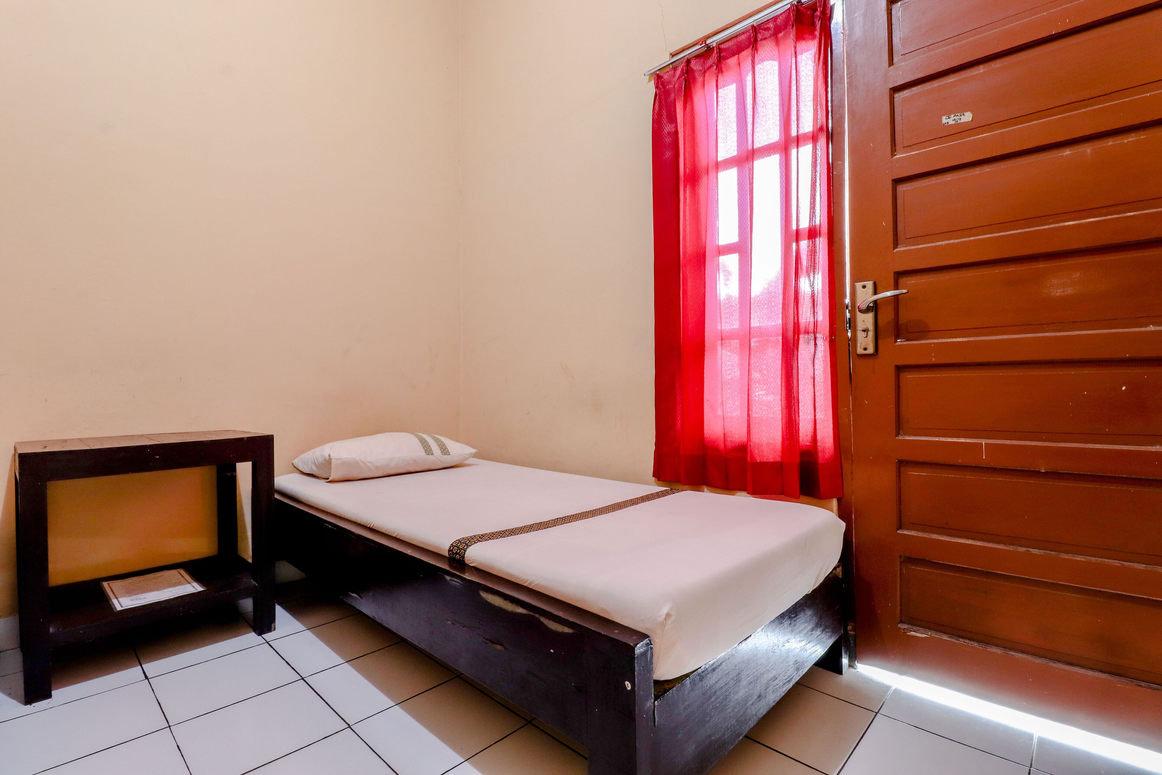 A small, single-bed room with a wooden bed frame, a nightstand, and a red curtain.