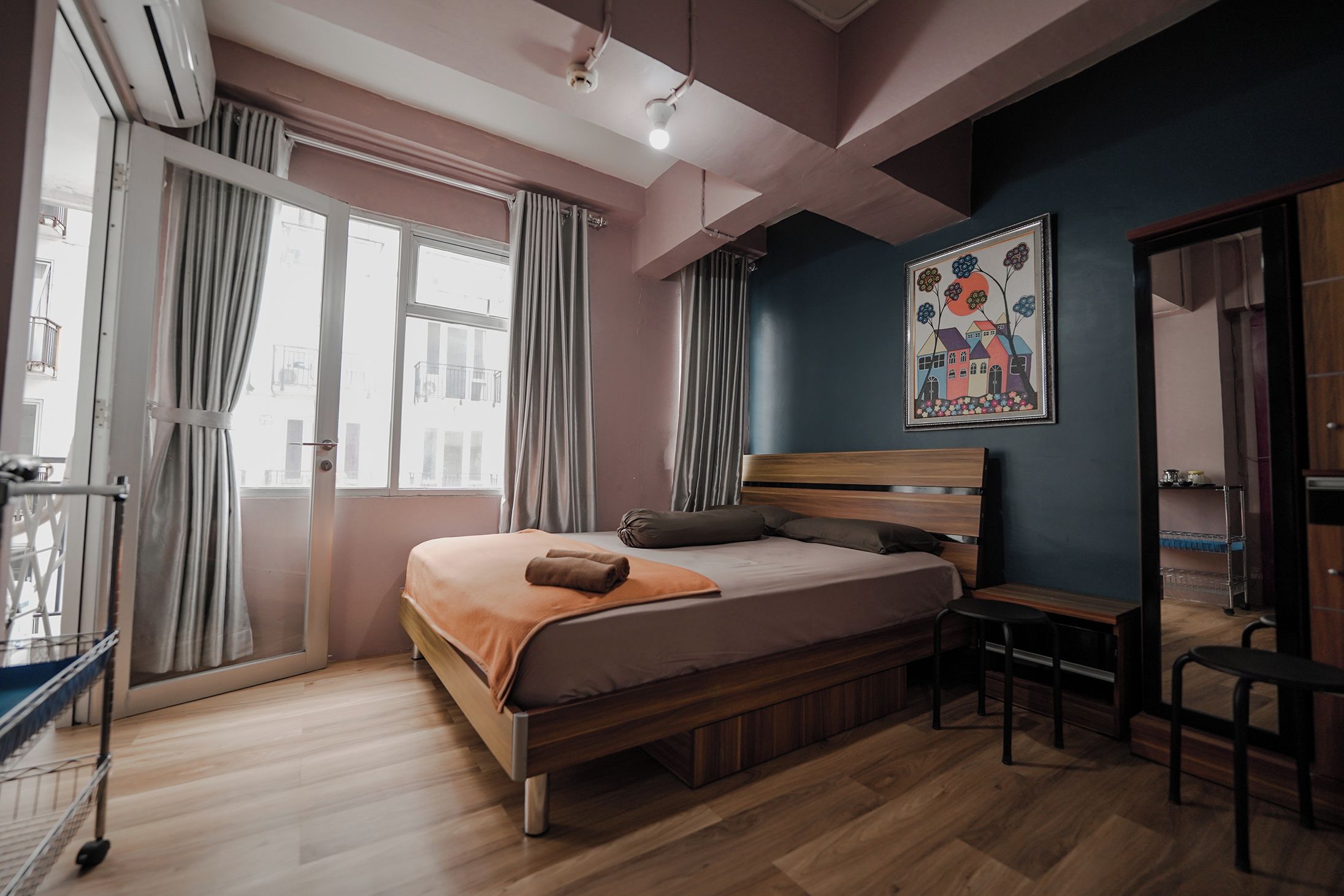 A bedroom with a bed, a desk, and a chair. There is a window with curtains, a painting on the wall, and an air conditioning unit.