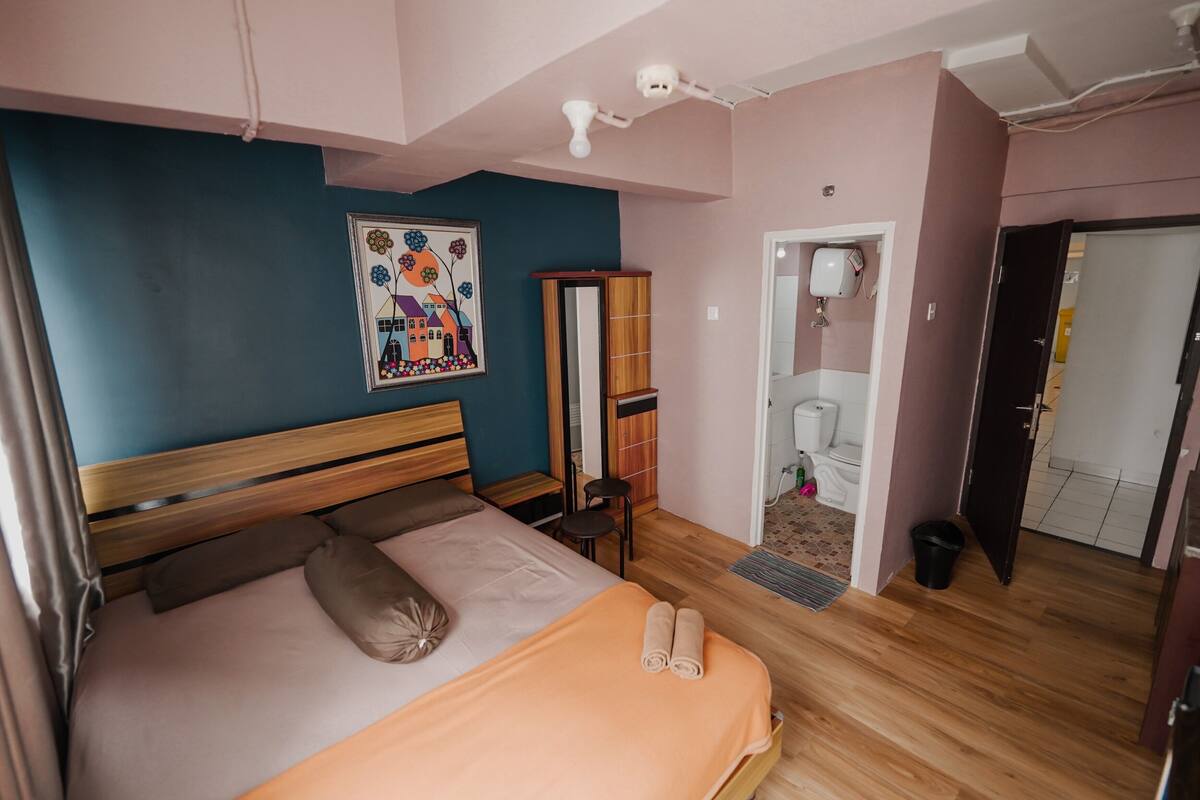 Un dormitorio con una cama, cabecera de madera, un cuadro, un espejo, una mesita, una silla, un inodoro y una puerta que da a otra habitación.