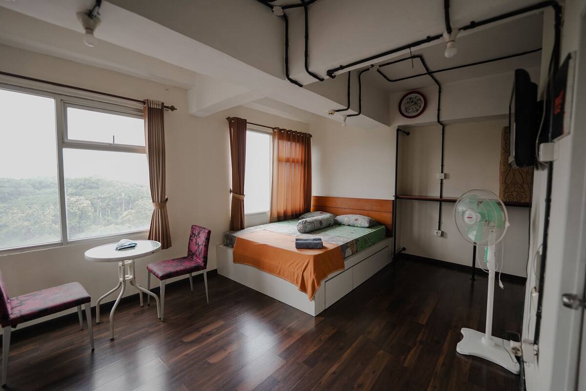 Un dormitorio con cama, una mesa, dos sillas, un ventilador de pie y una ventana con cortinas.