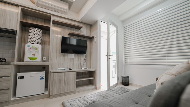 A modern living room with a grey sofa, a flat-screen TV mounted on a wooden wall, a small fridge, and a window with blinds. - The Jarrdin by Bethania Pro (Bandung)