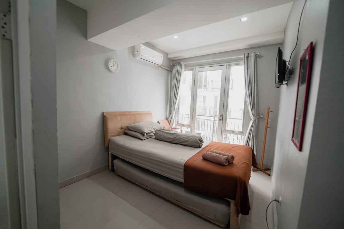 Un dormitorio con cama, ventana con cortinas, unidad de aire acondicionado y un reloj en la pared.