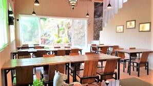 A restaurant with wooden tables and chairs, large windows with a view of greenery, and pendant lighting. - Anargya Guest House (Malang)