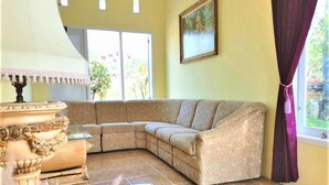 A living room with a beige sectional sofa, a decorative table, and a painting on the wall. - Anargya Guest House (Malang)
