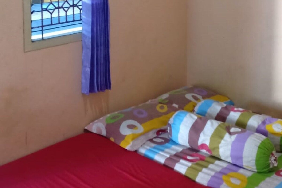 Una habitación pequeña con cama individual, colcha roja, almohadas coloridas y ventana con cortina azul.