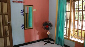 Una habitación con puerta de madera, un espejo, un ventilador de pie y una ventana con cortina.