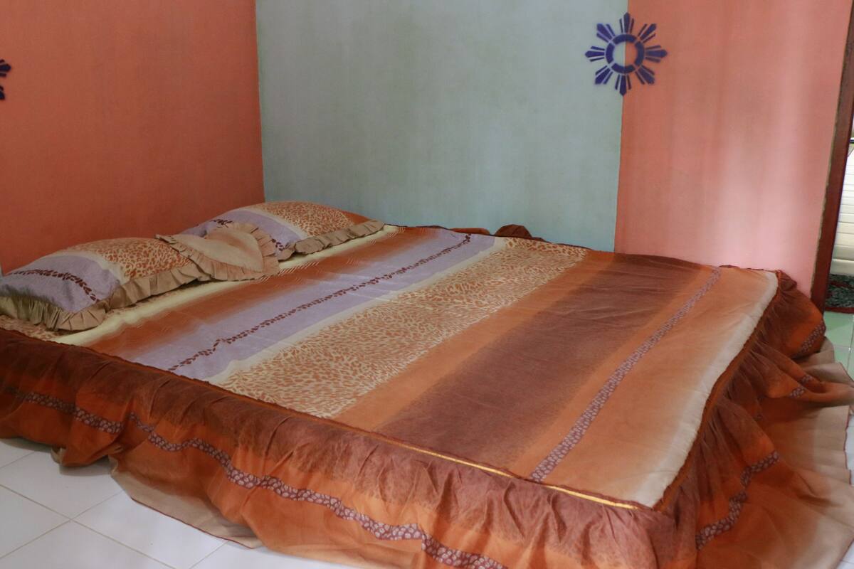A bed with a patterned bedspread and pillows, a wooden dresser, and a tiled floor.