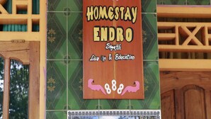 Un letrero con el texto 'HOMESTAY ENDRO' y 'Live & Education' en una tabla de madera, colgado en una pared con baldosas decorativas.