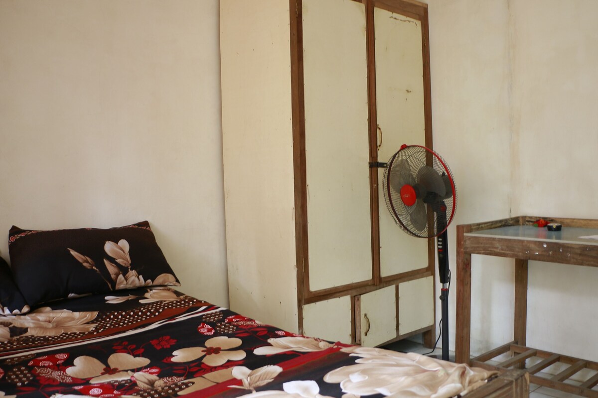 Ein Zimmer mit einem Bett, einem Holztisch und einem Standventilator.