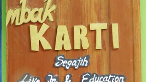 Un cartello di legno con il testo "HOMESTAY mbak KARTI" e "Segajih Live In & Education", arricchito da elementi decorativi.