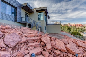 A modern two-story house with a stone retaining wall in front, a staircase leading to a balcony, and a view of red rock formations in the background.