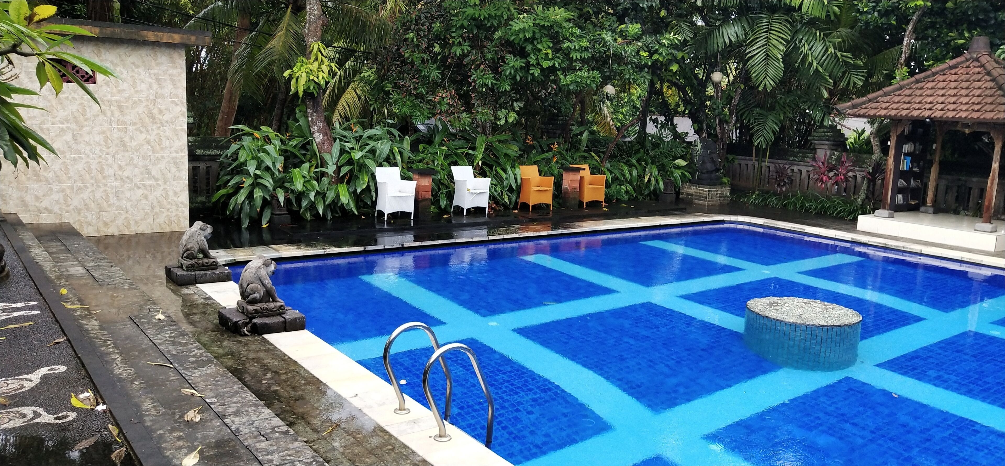 Una piscina con suelo de azulejos azules, rodeada de follaje y mobiliario de exterior.