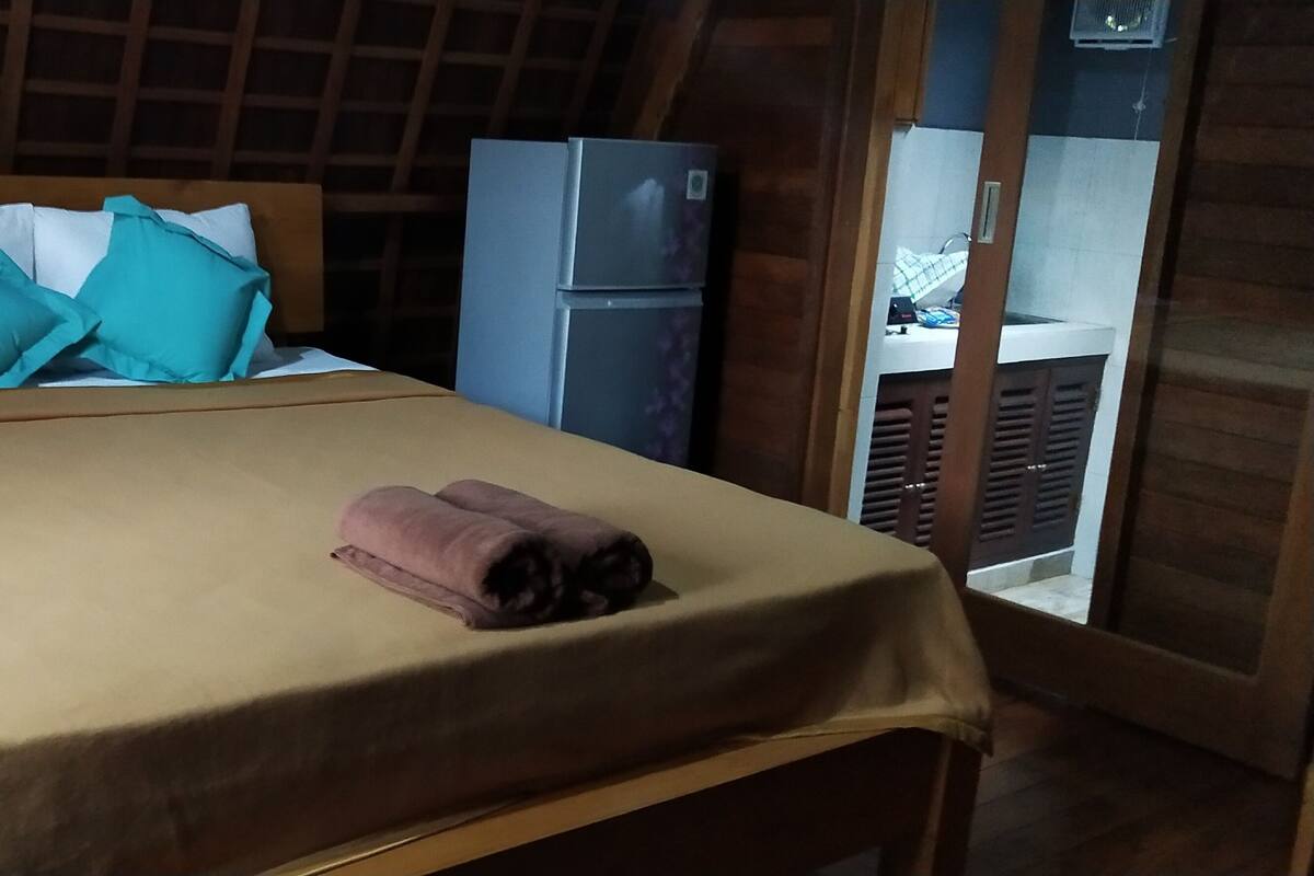 Quarto com cama, almofadas, toalha enrolada, mesa de cabeceira e geladeira.