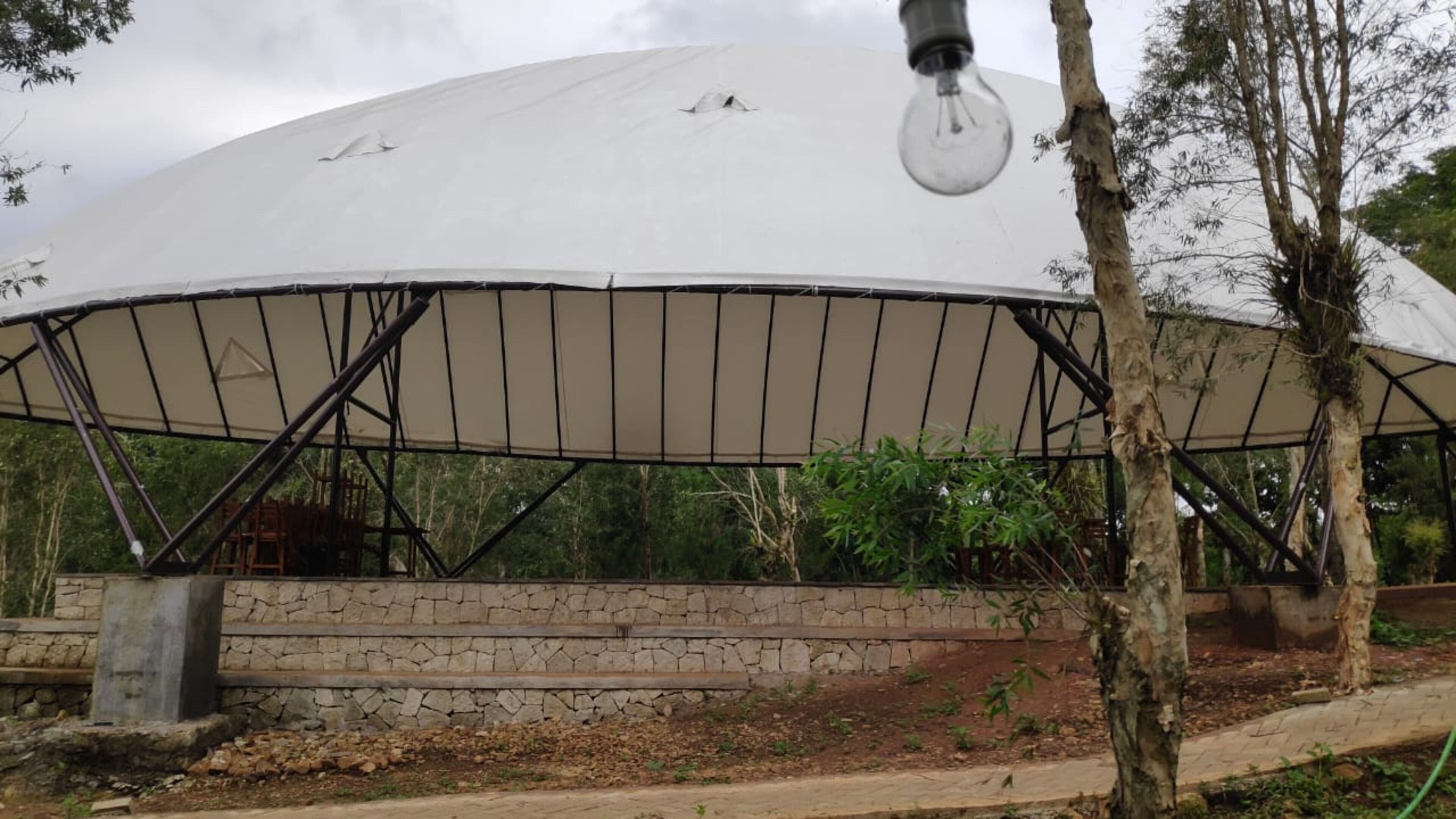 A large, dome-shaped pavilion with a stone base and metal framework, surrounded by trees and vegetation.