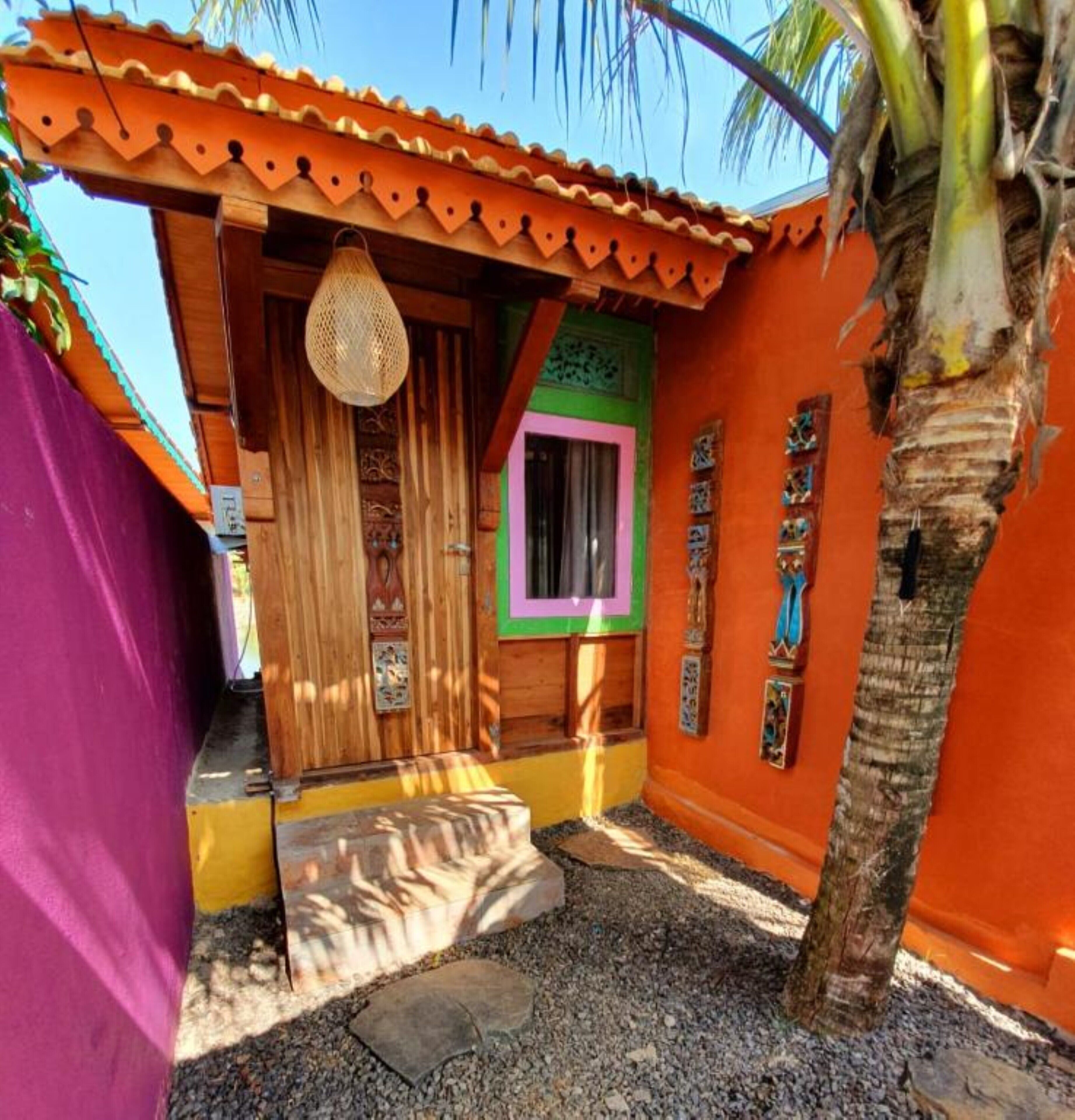 Un pequeño edificio colorido con una puerta de madera, una cesta tejida colgando sobre la puerta y un ventanal con marco rosa.