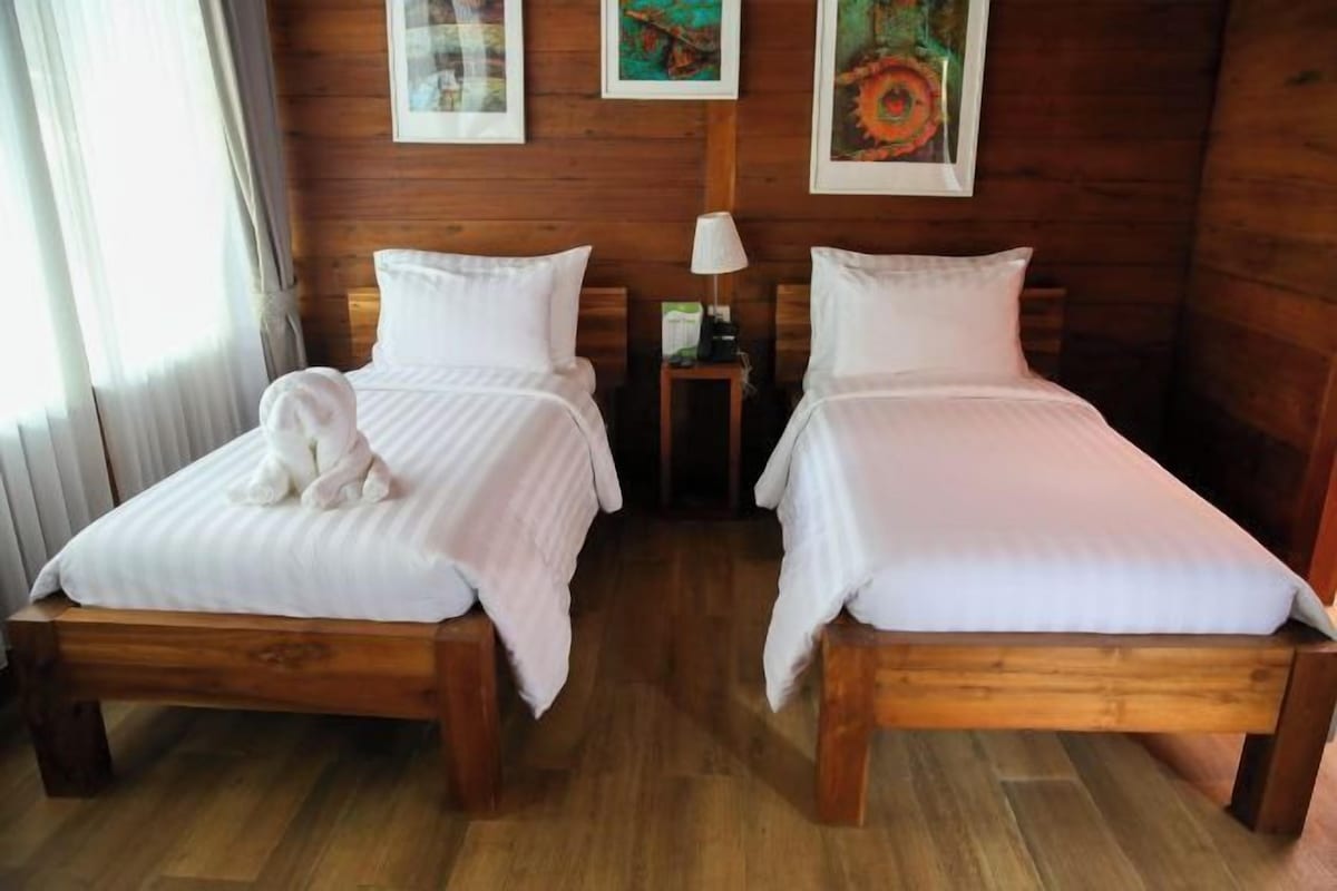 A room with two wooden beds, white bedding, a small table with a lamp, and artwork on the walls.