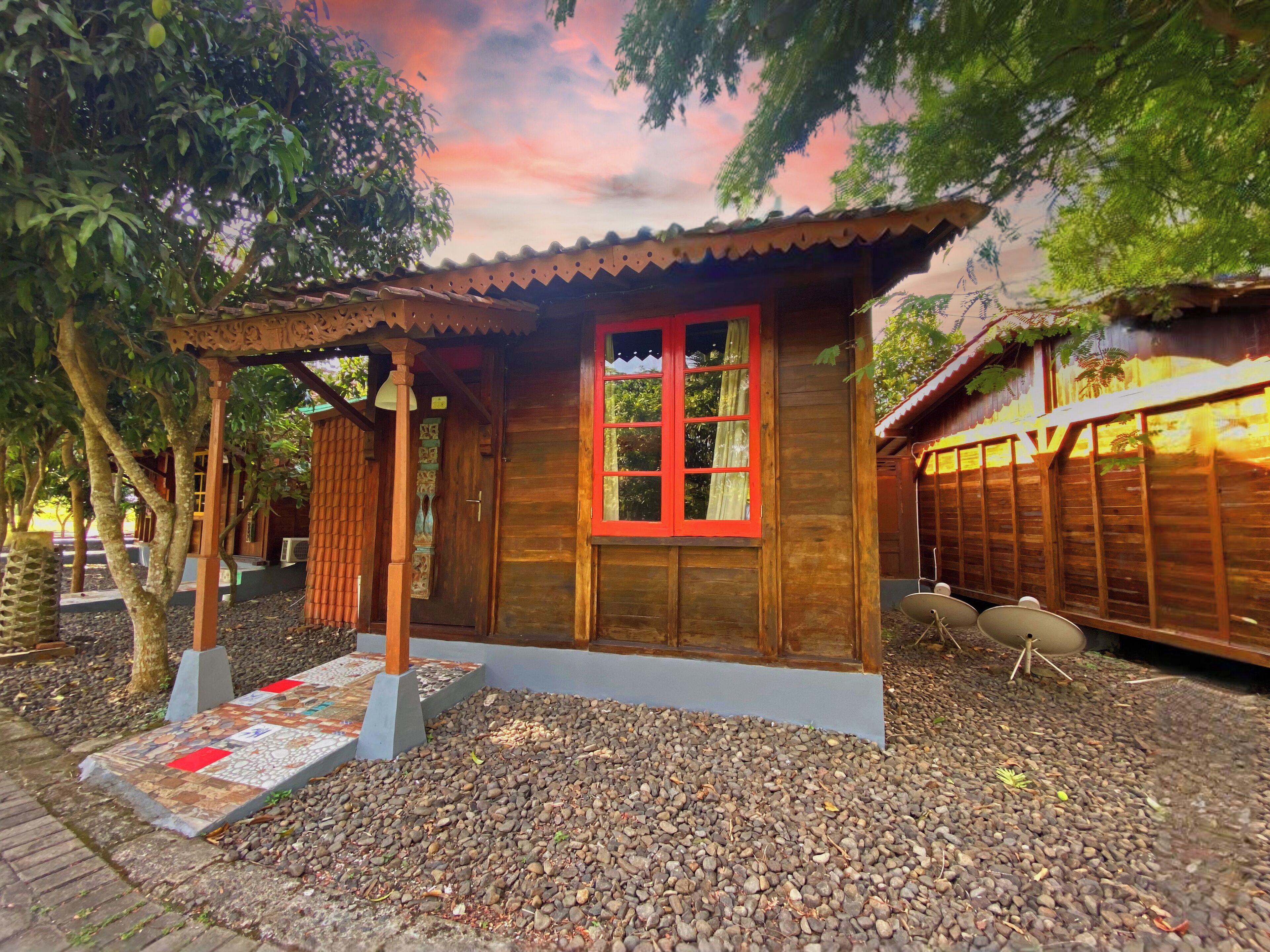 A wooden cabin with a red-framed window, surrounded by trees and a pebbled ground.