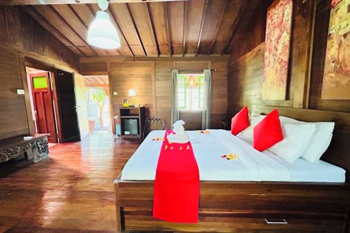 A room with a wooden floor, a bed with white bedding and red pillows, a small table with a lamp, and a painting on the wall.