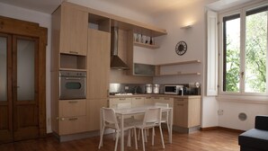 A modern kitchen with wooden cabinets, a built-in oven, a dining table with chairs, and a window with a view of greenery.