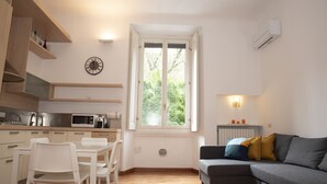 A modern kitchen with white cabinets, a dining table with chairs, a gray sofa with cushions, and a large window with a view of greenery.