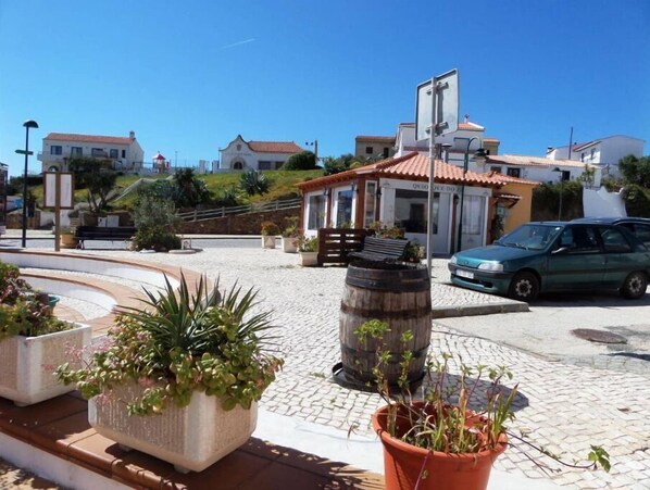 Una calle adoquinada con una pequeña plaza, un barril de madera y un coche verde estacionado al lado.